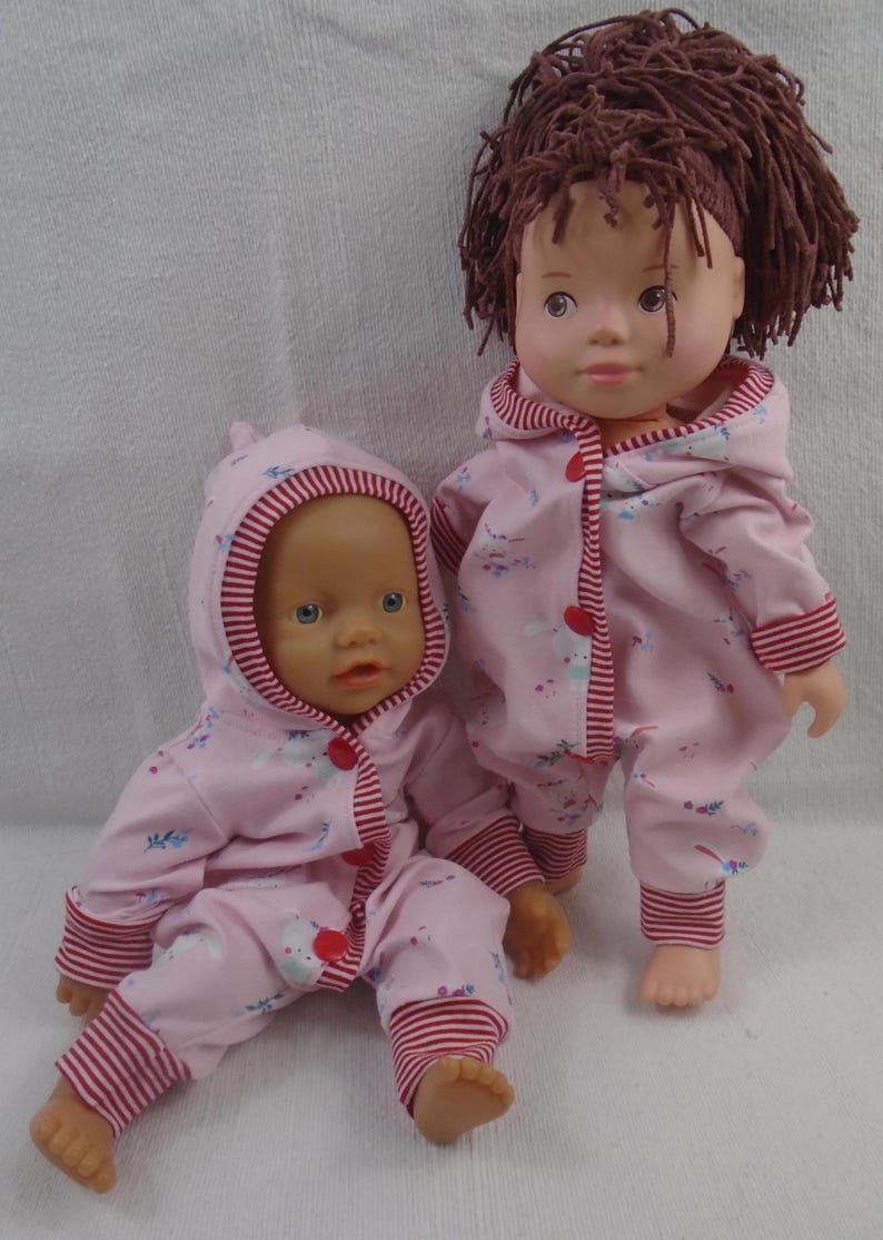 Puede incluir: Dos mu&ntilde;ecas con monos rosas con capucha a juego con ribetes a rayas rojas y blancas. Los monos tienen un peque&ntilde;o estampado floral y botones rojos. Una mu&ntilde;eca est&aacute; sentada, la otra de pie.