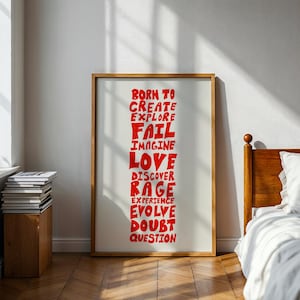 Peut inclure: Une affiche encadrée avec un fond blanc et du texte rouge. Le texte comprend des mots comme "BORN TO CREATE EXPLORE FAIL IMAGINE LOVE DISCOVER RAGE EXPERIENCE EVOLVE DOUBT QUESTION". Le cadre est de couleur marron clair.