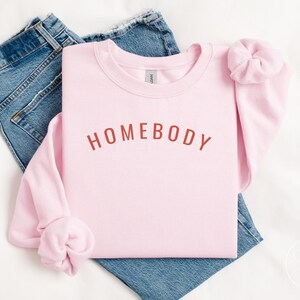 May include: A light pink sweatshirt with the word "HOMEBODY" in a rust-colored font. The sweatshirt is folded on top of a pair of blue jeans. A matching pink scrunchie is placed on the sleeve.