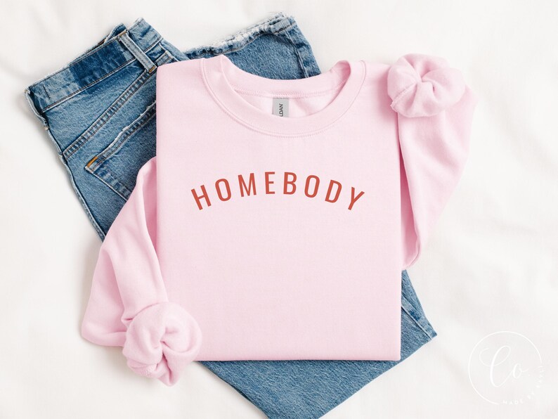 May include: A light pink sweatshirt with the word "HOMEBODY" in a rust-colored font. The sweatshirt is folded on top of a pair of blue jeans. A matching pink scrunchie is placed on the sleeve.