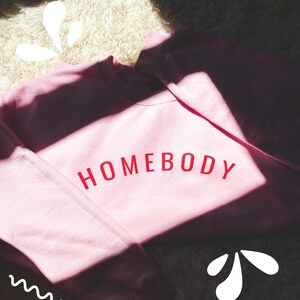 May include: A light pink sweatshirt with the word "HOMEBODY" printed in red across the chest. The sweatshirt is made of a soft material and is likely designed for comfort. The image is well-lit, with shadows adding depth.