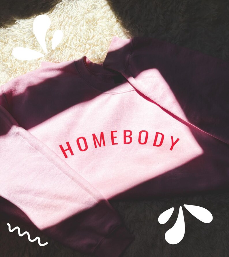 May include: A light pink sweatshirt with the word "HOMEBODY" printed in red across the chest. The sweatshirt is made of a soft material and is likely designed for comfort. The image is well-lit, with shadows adding depth.