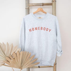 May include: A light gray sweatshirt with the word "HOMEBODY" in red lettering. The sweatshirt is hanging on a wooden hanger against a bamboo ladder. Dried decorative plants are in the foreground.