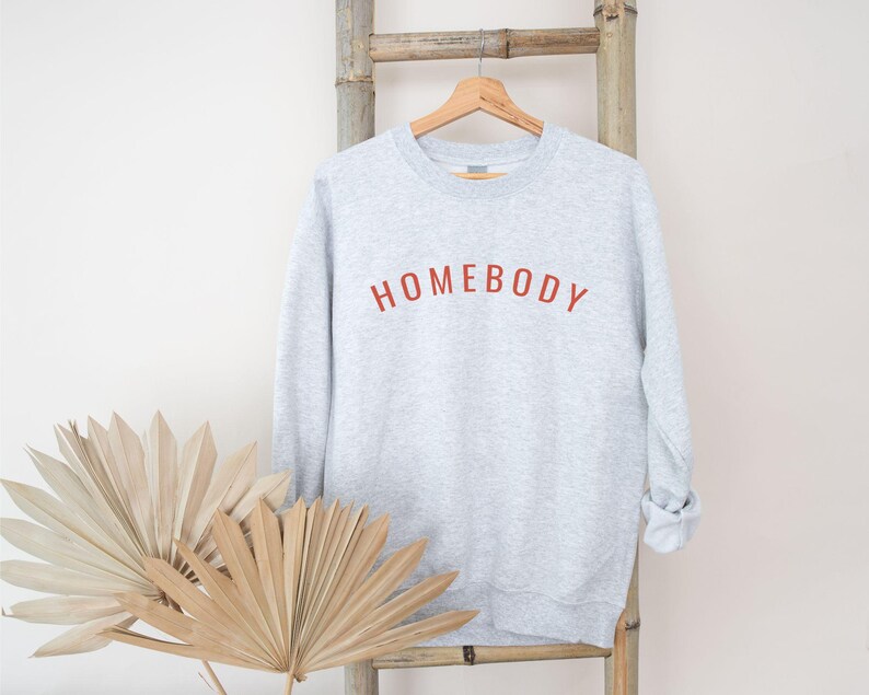 May include: A light gray sweatshirt with the word "HOMEBODY" in red lettering. The sweatshirt is hanging on a wooden hanger against a bamboo ladder. Dried decorative plants are in the foreground.