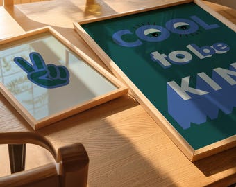Puede incluir: Dos impresiones enmarcadas sobre una mesa de madera. La impresión de la izquierda tiene un signo de paz azul. La impresión de la derecha tiene un fondo turquesa con el texto "COOL to be KIND" en azul y blanco.