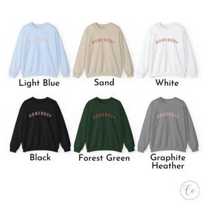 May include: Six crewneck sweatshirts in various colors: light blue, sand, white, black, forest green, and graphite heather. Each sweatshirt has the word "HOMEBODY" printed across the chest in a contrasting color. The sweatshirts are displayed on a white background.