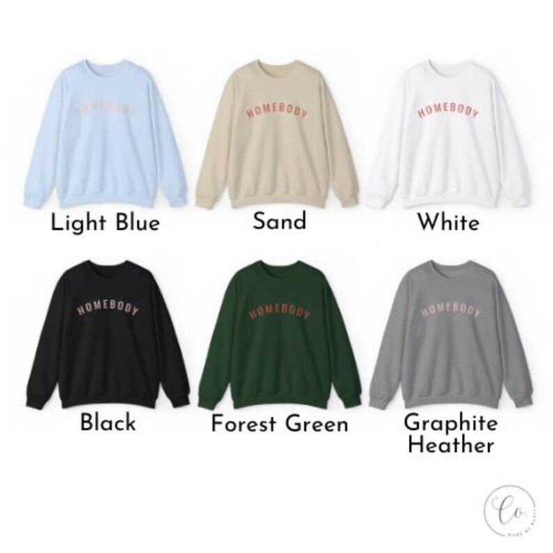 May include: Six crewneck sweatshirts in various colors: light blue, sand, white, black, forest green, and graphite heather. Each sweatshirt has the word "HOMEBODY" printed across the chest in a contrasting color. The sweatshirts are displayed on a white background.