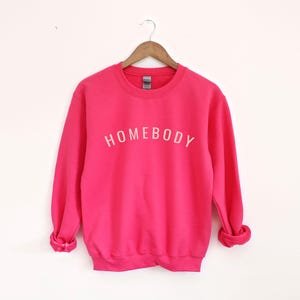 May include: A bright pink sweatshirt with the word "HOMEBODY" printed in white across the chest. The long-sleeved crew neck top is displayed on a wooden hanger against a white background. The sweatshirt is a solid color.
