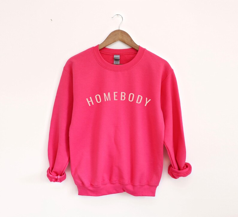 May include: A bright pink sweatshirt with the word "HOMEBODY" printed in white across the chest. The long-sleeved crew neck top is displayed on a wooden hanger against a white background. The sweatshirt is a solid color.