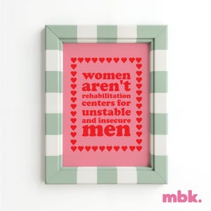 May include: A framed print with a pink background and a white and green checkered frame. The print features the text "women aren't rehabilitation centers for unstable and insecure men" in red letters surrounded by a border of red hearts.