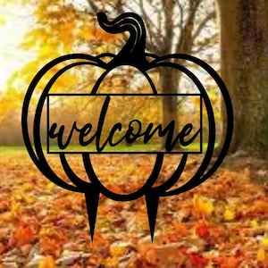 May include: Black metal pumpkin silhouette with the word "welcome" in a banner across the middle. The pumpkin is set against a background of autumn leaves.