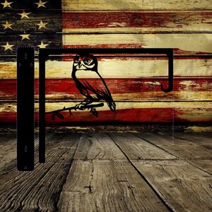 May include: Black metal hanging bracket featuring a silhouette of an owl perched on a branch. The bracket is against a backdrop of the American flag on a rustic wooden surface. The design is suitable for outdoor or indoor use.