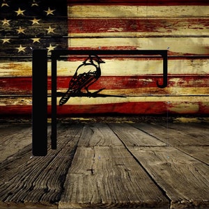 May include: Black metal flag hanger with a silhouette of a blue jay perched on a branch. The hanger is mounted against a weathered wood background with an American flag.