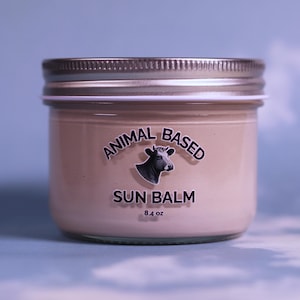 Puede incluir: Un frasco de vidrio con tapa de metal plateado que contiene un bálsamo beige. La etiqueta presenta una ilustración de una vaca en blanco y negro con el texto ANIMAL BASED SUN BALM y 8.4 oz impreso en negro sobre el envase claro.