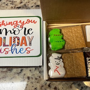 May include: A festive s'mores kit in a white box with a green border. The lid says "Wishing you S'more Holiday Wishes." Inside are graham crackers, chocolate bars, snowman and Christmas tree marshmallows, a candle, and matches for a treat.