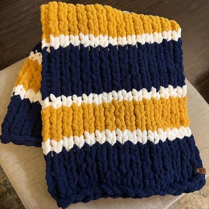 May include: A striped knitted throw blanket in navy blue, white, and mustard yellow. The blanket is made with chunky yarn and has a soft, fluffy texture.