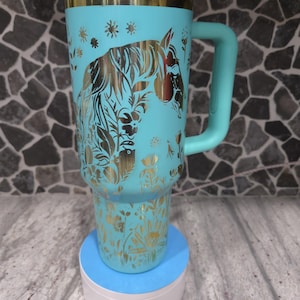 Pu&ograve; includere: Una tazza da viaggio turchese con un design di cavallo e fiori dorati. La tazza ha un coperchio in acciaio inossidabile e un manico.