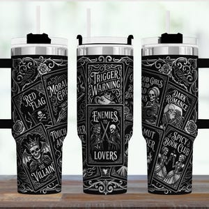 May include: Black and white insulated tumbler with a handle, featuring a gothic design. The tumbler is covered in illustrations of playing cards with phrases like "Trigger Warning", "Enemies to Lovers", and "Spicy Book Club". It includes a lid and a straw.