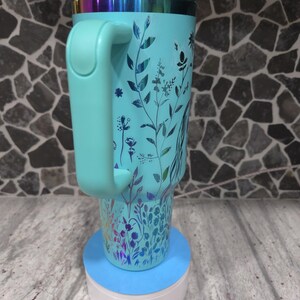 Pu&ograve; includere: Una tazza da viaggio turchese con un motivo floreale e un coperchio abbinato. La tazza ha un manico e si trova su un sottobicchiere blu e bianco.