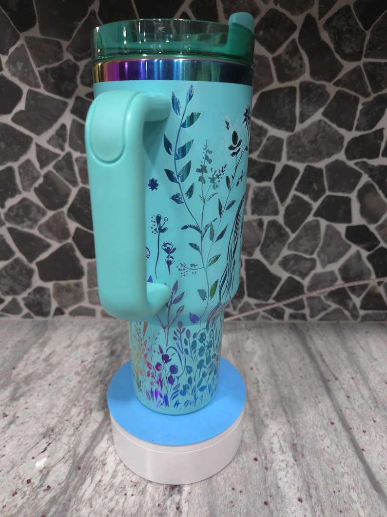 Pu&ograve; includere: Una tazza da viaggio turchese con un motivo floreale e un coperchio abbinato. La tazza ha un manico e si trova su un sottobicchiere blu e bianco.