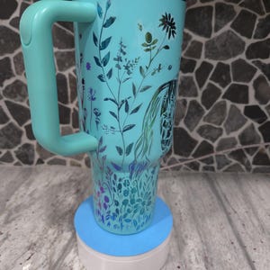 Pu&ograve; includere: Un bicchiere in acciaio inossidabile blu turchese con un motivo floreale e un manico abbinato. Il bicchiere &egrave; appoggiato su un sottobicchiere blu e bianco.