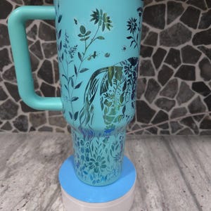 Pu&ograve; includere: Una tazza da viaggio blu turchese con un motivo floreale e un coperchio verde. La tazza ha un manico ed &egrave; appoggiata su un sottobicchiere blu e bianco.