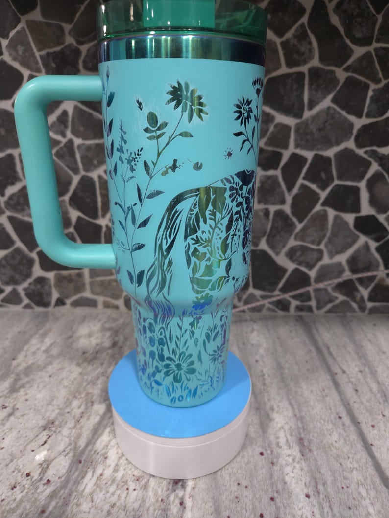 Pu&ograve; includere: Una tazza da viaggio blu turchese con un motivo floreale e un coperchio verde. La tazza ha un manico ed &egrave; appoggiata su un sottobicchiere blu e bianco.