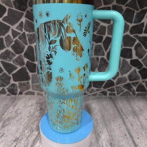 Pu&ograve; includere: Una tazza da viaggio turchese con un design di cavallo dorato e fiori. La tazza ha un coperchio verde e un manico. &Egrave; appoggiata su un sottobicchiere blu e bianco.