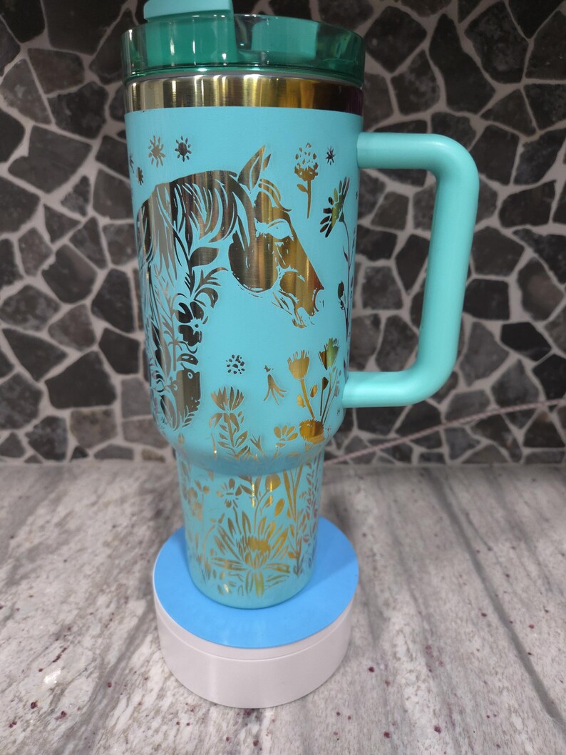 Pu&ograve; includere: Una tazza da viaggio turchese con un design di cavallo dorato e fiori. La tazza ha un coperchio verde e un manico. &Egrave; appoggiata su un sottobicchiere blu e bianco.