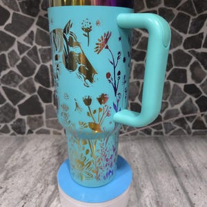 Pu&ograve; includere: Una tazza da viaggio turchese con un design floreale dorato e una silhouette di testa di cavallo. La tazza ha un coperchio e un manico. &Egrave; appoggiata su un sottobicchiere blu e bianco.
