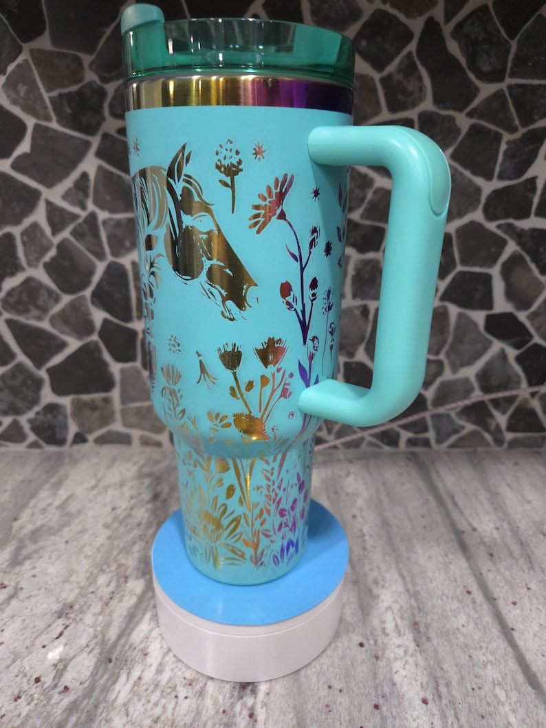 Pu&ograve; includere: Una tazza da viaggio turchese con un design floreale dorato e una silhouette di testa di cavallo. La tazza ha un coperchio e un manico. &Egrave; appoggiata su un sottobicchiere blu e bianco.