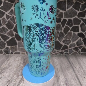 Pu&ograve; includere: Una tazza da viaggio turchese con un design floreale ed elefante. La tazza ha un manico ed &egrave; appoggiata su un sottobicchiere blu e bianco.