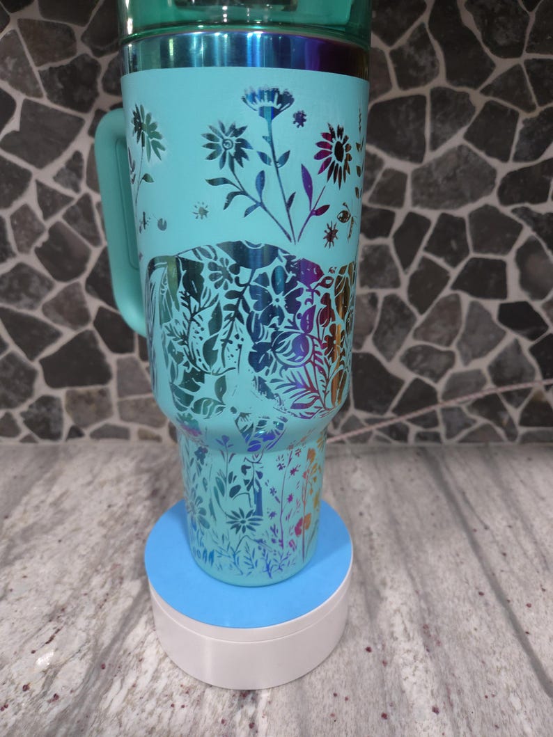 Pu&ograve; includere: Una tazza da viaggio turchese con un design floreale ed elefante. La tazza ha un manico ed &egrave; appoggiata su un sottobicchiere blu e bianco.