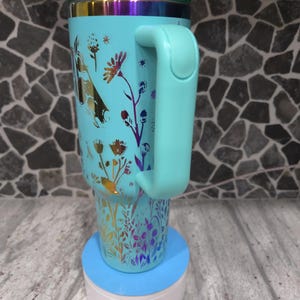 Pu&ograve; includere: Una tazza da viaggio blu turchese con un motivo floreale e un manico abbinato. La tazza &egrave; appoggiata su un sottobicchiere blu e bianco.