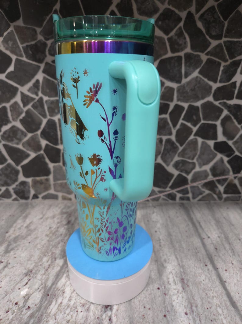 Pu&ograve; includere: Una tazza da viaggio blu turchese con un motivo floreale e un manico abbinato. La tazza &egrave; appoggiata su un sottobicchiere blu e bianco.