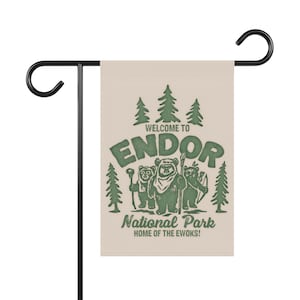 Puede incluir: Una bandera de jardín beige con un soporte de metal negro. La bandera presenta un gráfico verde con el texto "WELCOME TO ENDOR National Park HOME OF THE EWOKS!" e ilustraciones de Ewoks y árboles.