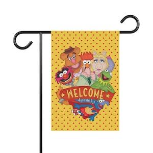 May include: A yellow garden flag with a red polka dot pattern featuring Muppet characters. The flag has a banner that reads "WELCOME friends". The flag is attached to a black metal garden stake.
