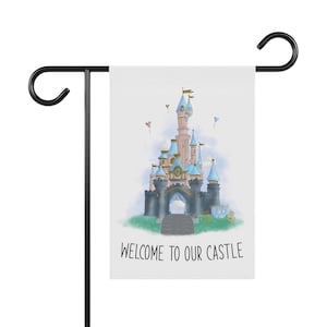 May include: A white garden flag with a cartoon illustration of a pink and blue castle with a gold clock tower. The castle is surrounded by clouds and there are two pink balloons in the sky. The text "WELCOME TO OUR CASTLE" is printed below the castle.