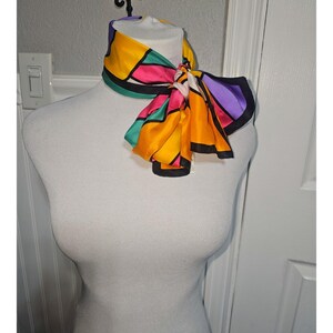 Puede incluir: Un pañuelo de seda colorido con un diseño geométrico. El pañuelo presenta una vibrante mezcla de colores, incluyendo amarillo, rosa, verde, naranja y morado, delineados en negro. El pañuelo está anudado, mostrando su audaz patrón.