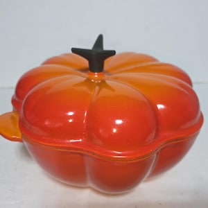 Pode incluir: Uma travessa de cerâmica brilhante em forma de abóbora laranja com uma haste preta. A travessa tem tampa e pega. O gradiente de cor vai do vermelho escuro ao laranja brilhante. Um item de cozinha decorativo.