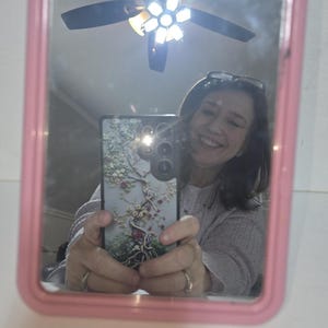 May include: A pink framed mirror reflecting a person holding a phone with a floral design case. The mirror also reflects a ceiling fan and a light fixture. The person is smiling.
