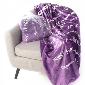 May include: Purple fleece throw blanket with white text that says "Hugs!" and "Warm Hugs" on a gray armchair.