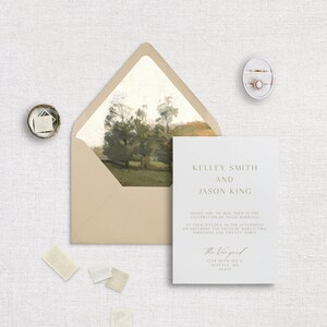 May include: Wedding invitation suite featuring a cream-colored invitation with gold text, an envelope with a landscape painting inside, and two rings in a white box. The invitation reads "Kelley Smith and Jason King" and includes the wedding details.