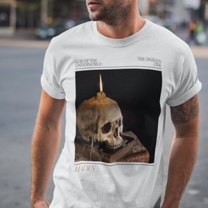 May include: White t-shirt with a graphic of a skull with a candle on top. The text "God of the Underworld" and "The Unseen One" are above the graphic. The text "Hades" is below the graphic.