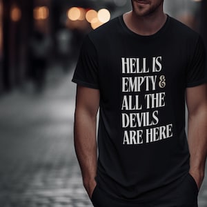 Puede incluir: Camiseta negra con el texto blanco "HELL IS EMPTY & ALL THE DEVILS ARE HERE." La camiseta es de cuello redondo y parece estar hecha de un material suave. La persona lleva pantalones negros.