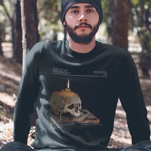 May include: A person wearing a black sweatshirt with a skull graphic and the text "God of the Underworld" and "The Singlen One". The person is sitting in a forest setting.