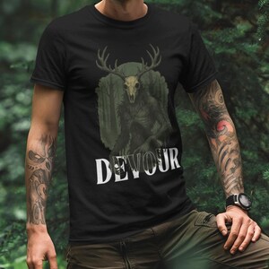 以下が含まれることがあります： 黒いTシャツ。角のある頭蓋骨のグラフィックと白い文字で「DEVOUR」と書かれています。デザインは森の背景に設定されています。Tシャツは森の中で人が着ています。