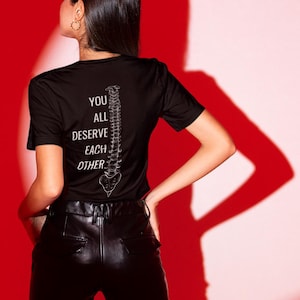May include: Black t-shirt with the text "YOU ALL DESERVE EACH OTHER" and a white spine graphic. The model is wearing black leather trousers. The background is red.