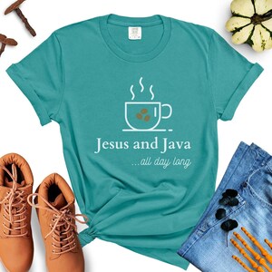 以下が含まれることがあります： 白のコーヒーカップのグラフィックと「Jesus and Java... all day long」というテキストが書かれたターコイズブルーのTシャツ。