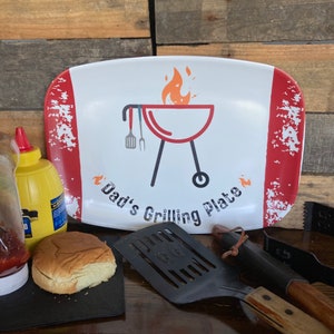May include: White ceramic serving platter with red and white distressed edges. The platter features a red and black grill with flames and the text "Dad's Grilling Plate".